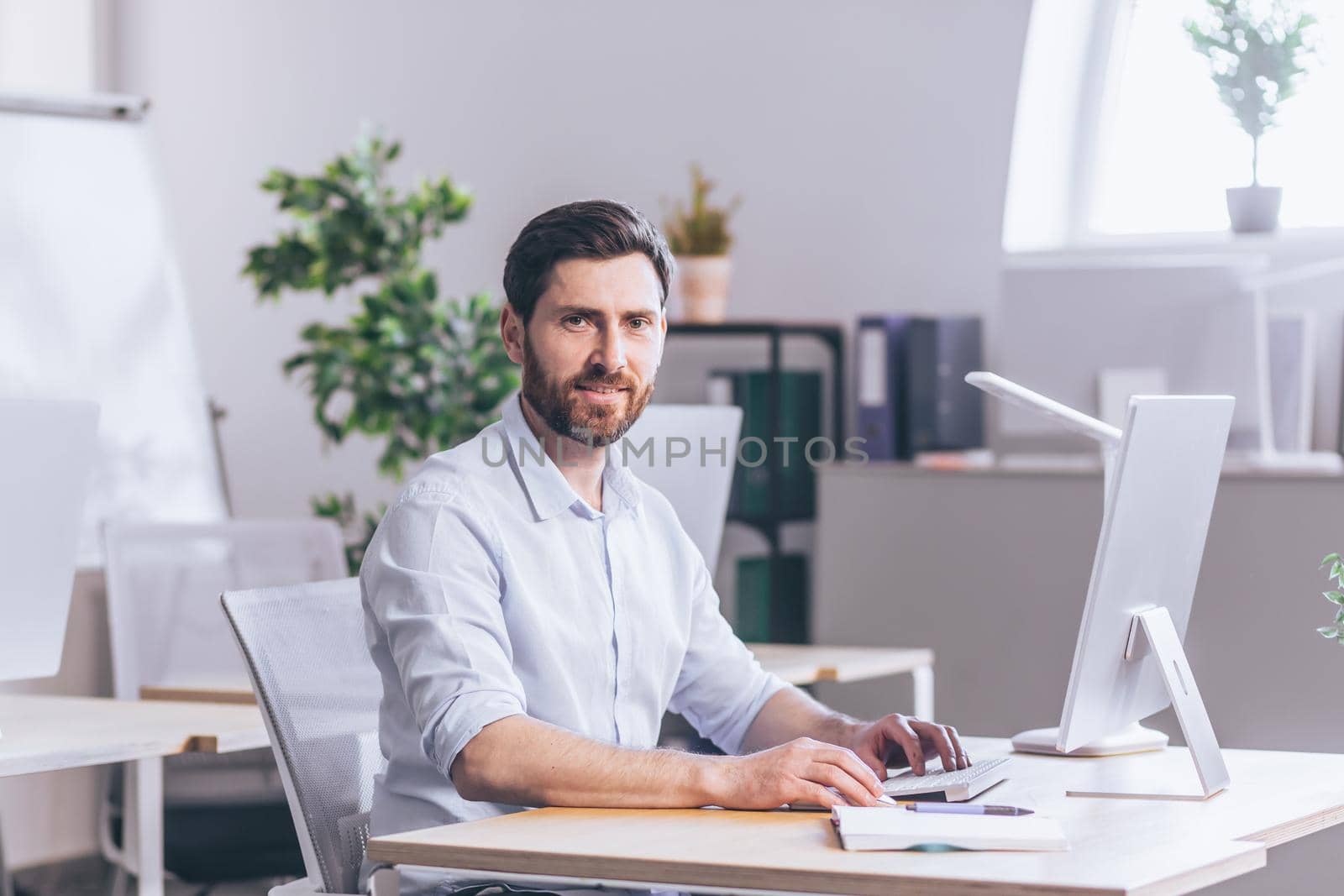 Successful bearded freelance businessman looks at camera and smiles, portrait of broker in office works at computer