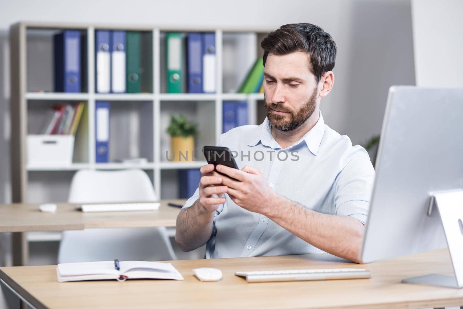Focused young man with a beard, uses the phone, sitting at home at the table, thoughtfully reads from the screen