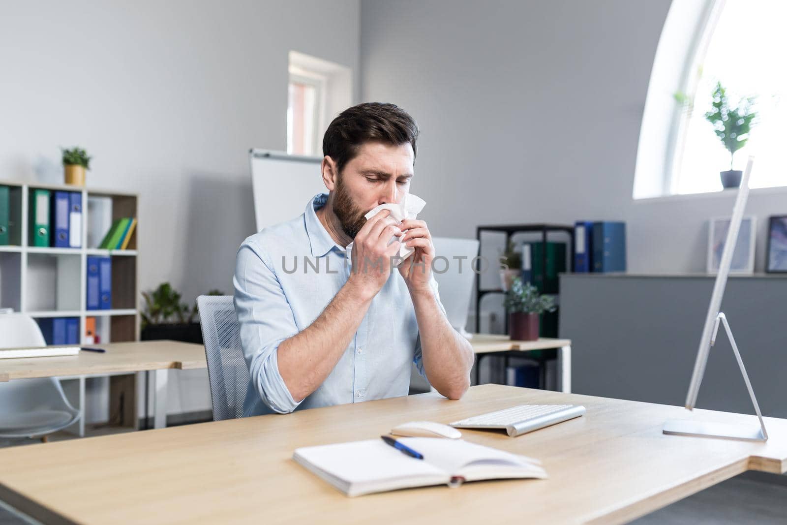 Businessman sick in the office man with allergies sneezes, sitting at a table with a computer by voronaman
