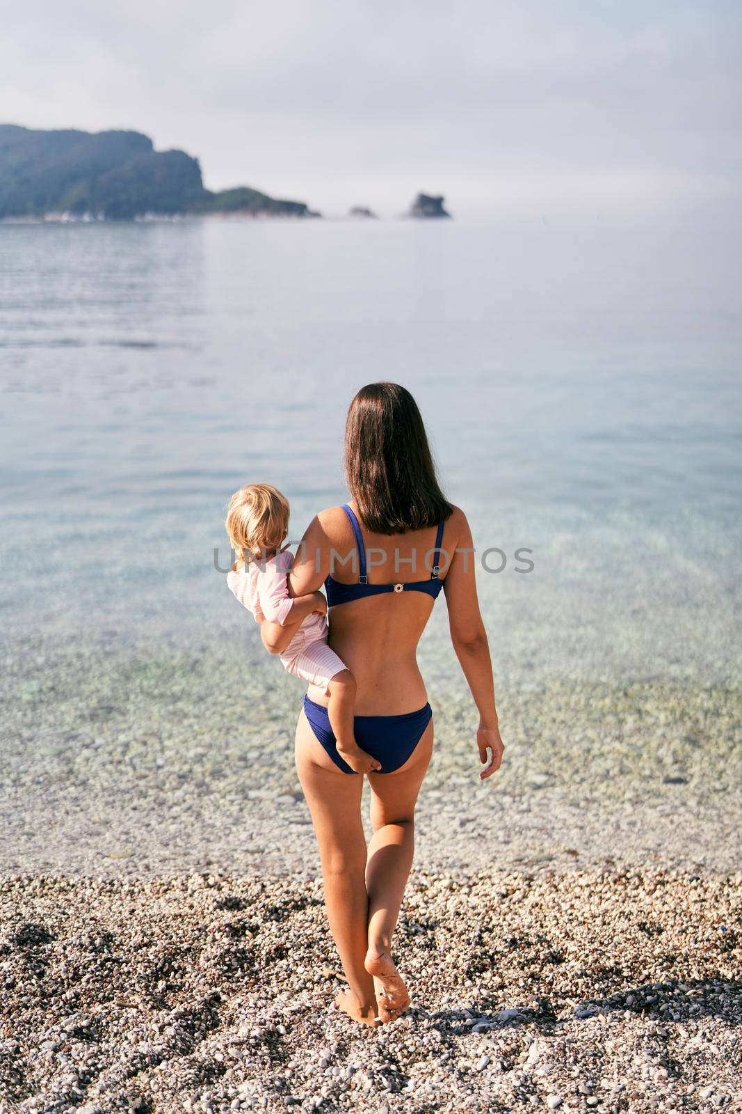 Mom with a little girl in her arms walks along a pebble beach to the sea. High quality photo