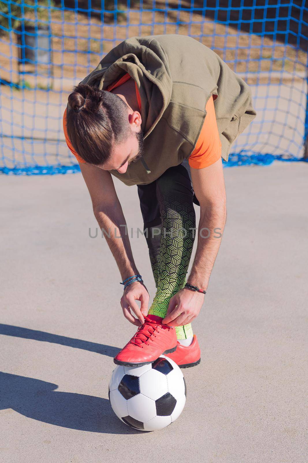 vertical photo of a football player tying his boots with a foot on the ball in a concrete soccer field, concept of healthy lifestyle and urban sport in the city, copy space for text