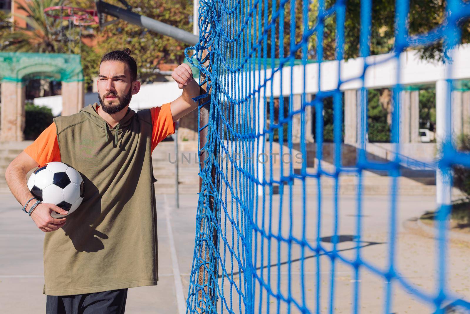 portrait of a handsome hipster soccer player leaning on the post next to the goal net with the ball under his arm, concept of healthy lifestyle and urban sport in the city, copy space for text