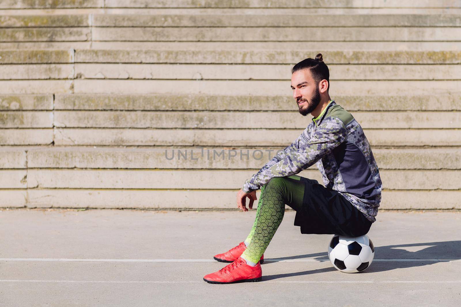 handsome sportsman with hair in a bun in a concrete soccer court sitting on his football ball, concept of healthy lifestyle and urban sport in the city, copy space for text