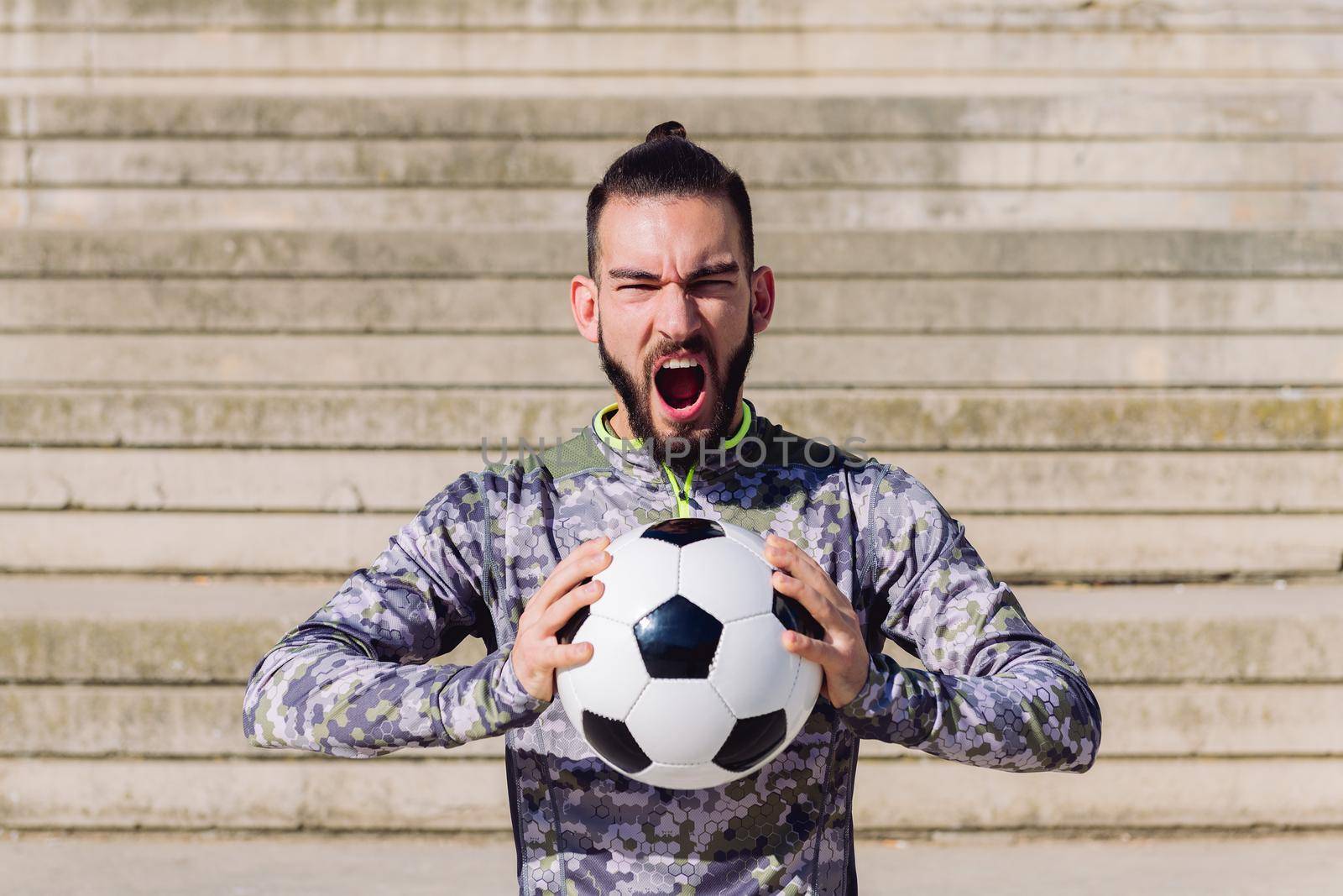 horizontal portrait of a handsome sportsman screaming in a concrete soccer court with the football ball in his hands, concept of healthy lifestyle and urban sport in the city, copy space for text
