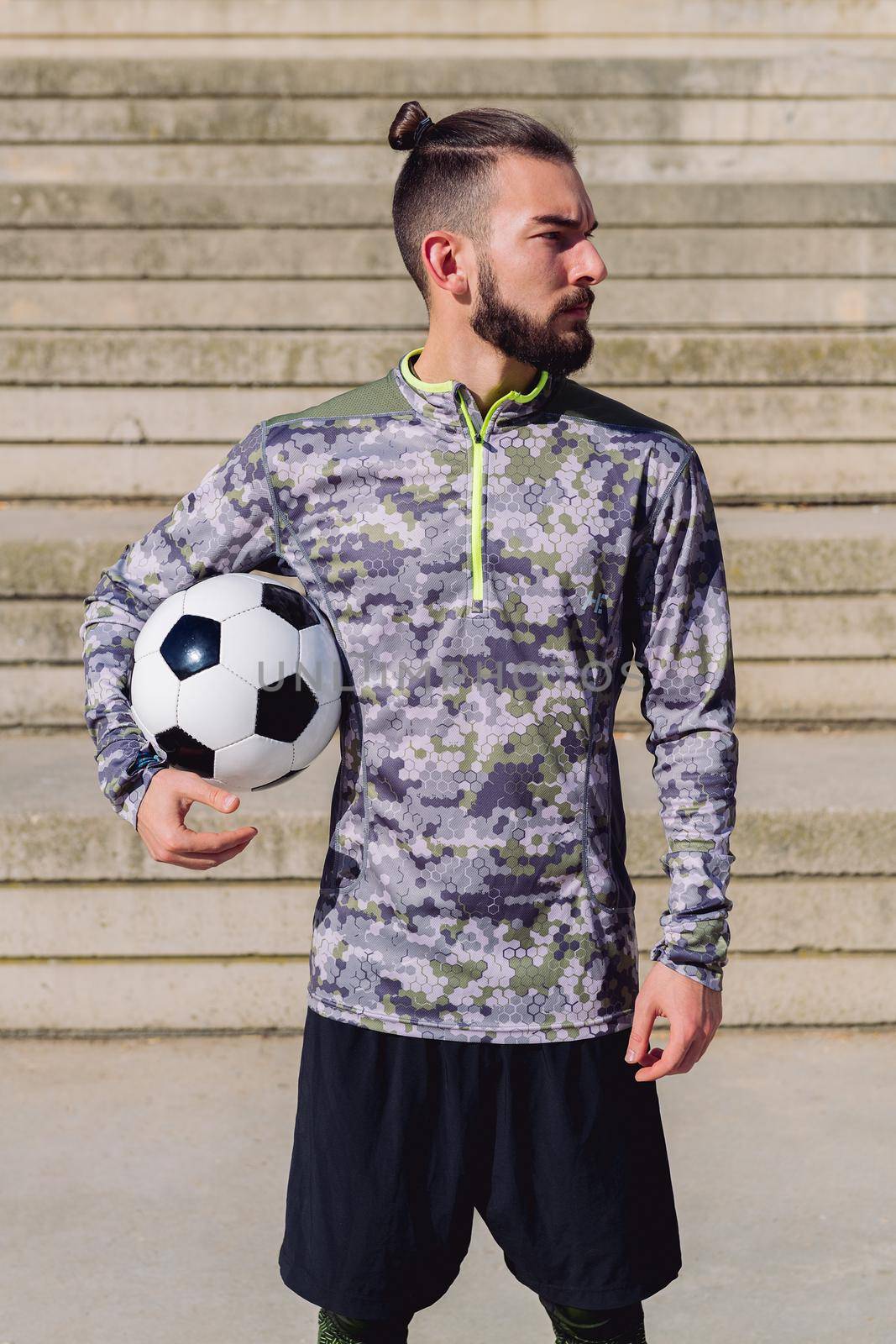 vertical photo of an attractive hipster sportsman in a concrete soccer court with the football ball under his arm, concept of healthy lifestyle and urban sport in the city