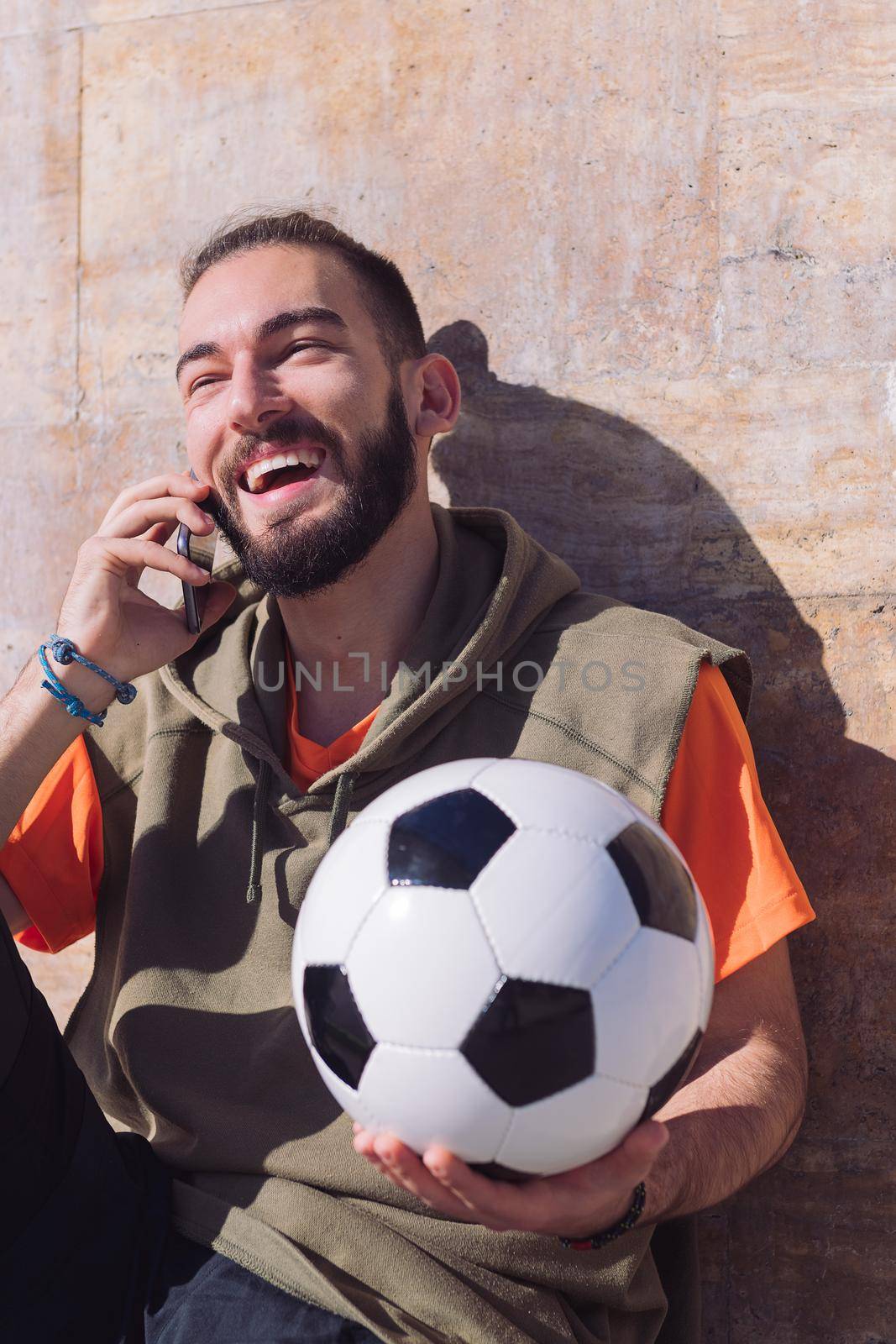 handsome sportsman laughing and talking by phone while resting with his soccer ball, concept of technology and urban sport lifestyle in the city, copy space for text