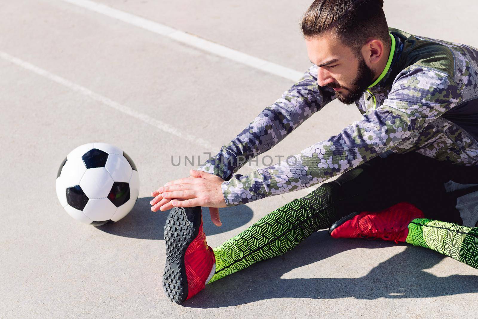 young footballer stretching legs sitting on the floor next to his football ball in a concrete soccer court, concept of healthy lifestyle and urban sport in the city, copy space for text