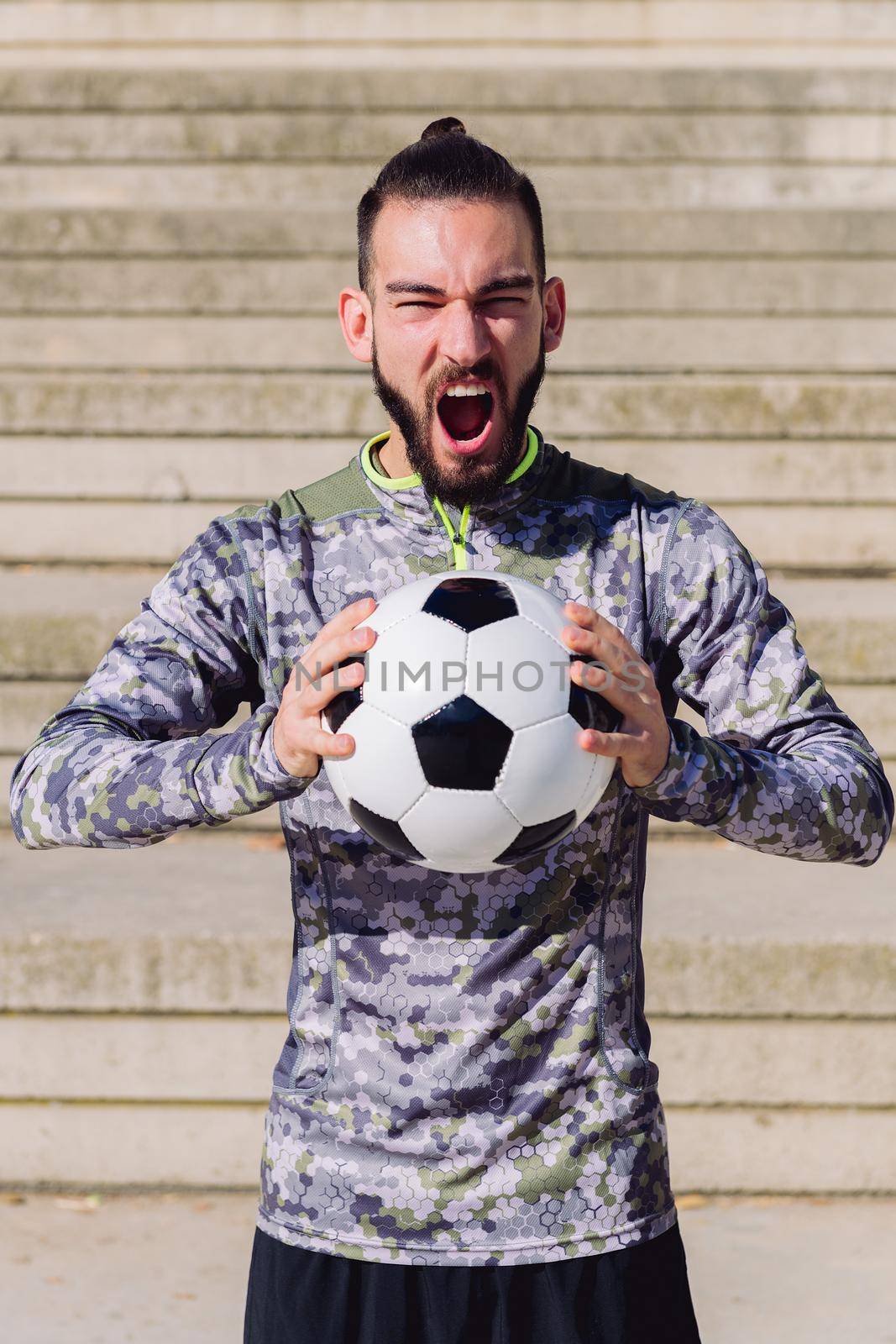 vertical portrait of a handsome sportsman screaming in a concrete soccer court with the football ball in his hands, concept of healthy lifestyle and urban sport in the city