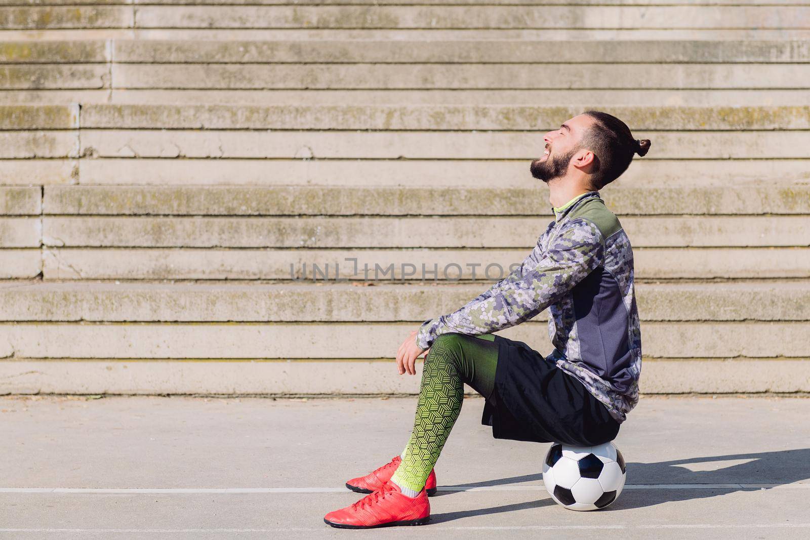 handsome sportsman with hair in a bun laughing in a concrete soccer court sitting on his football ball, concept of healthy lifestyle and urban sport in the city, copy space for text