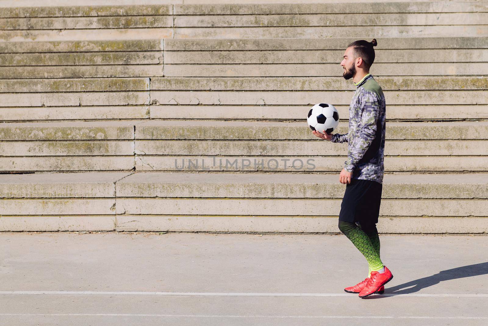 handsome sportsman with hair in a bun walking with the ball in his hand on a concrete soccer court, concept of healthy lifestyle and urban sport in the city, copy space for text