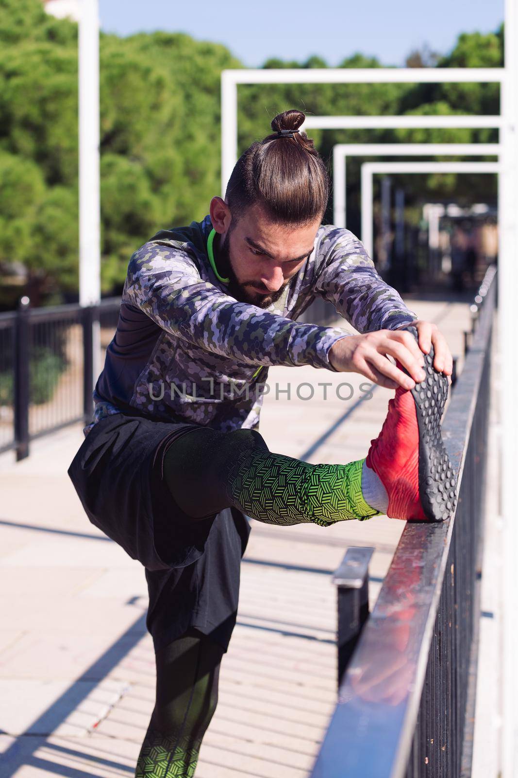 vertical photo of a handsome sportsman man with hair in a bun stretching the leg on a railing, concept of healthy lifestyle and urban sport in the city, copy space for text