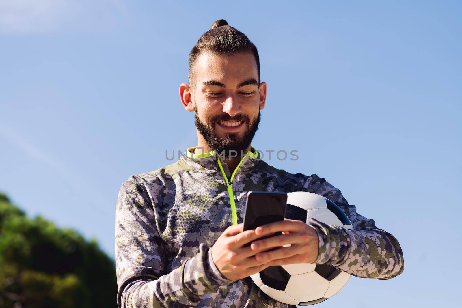 horizontal portrait of a happy sportsman with a soccer ball smiling writing a message on his mobile phone, concept of technology and urban sport lifestyle in the city, copy space for text