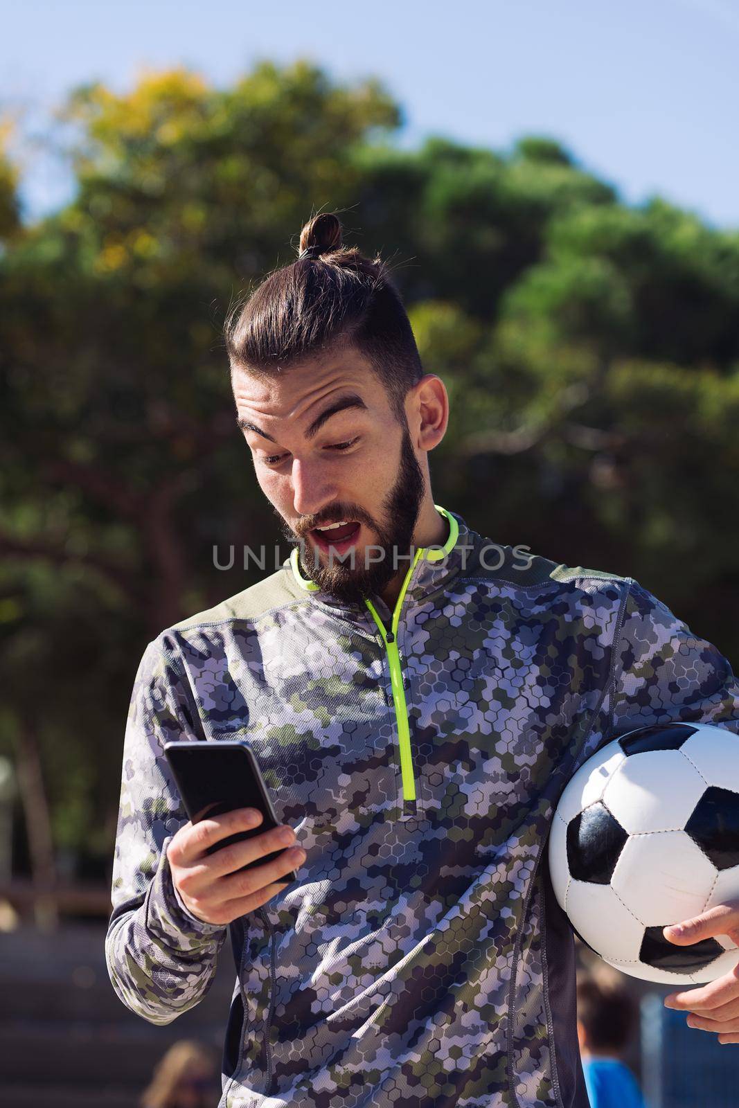 fashionable sportsman with a soccer ball consulting his mobile phone with surprised expression on the football court, concept of technology and urban sport lifestyle in the city