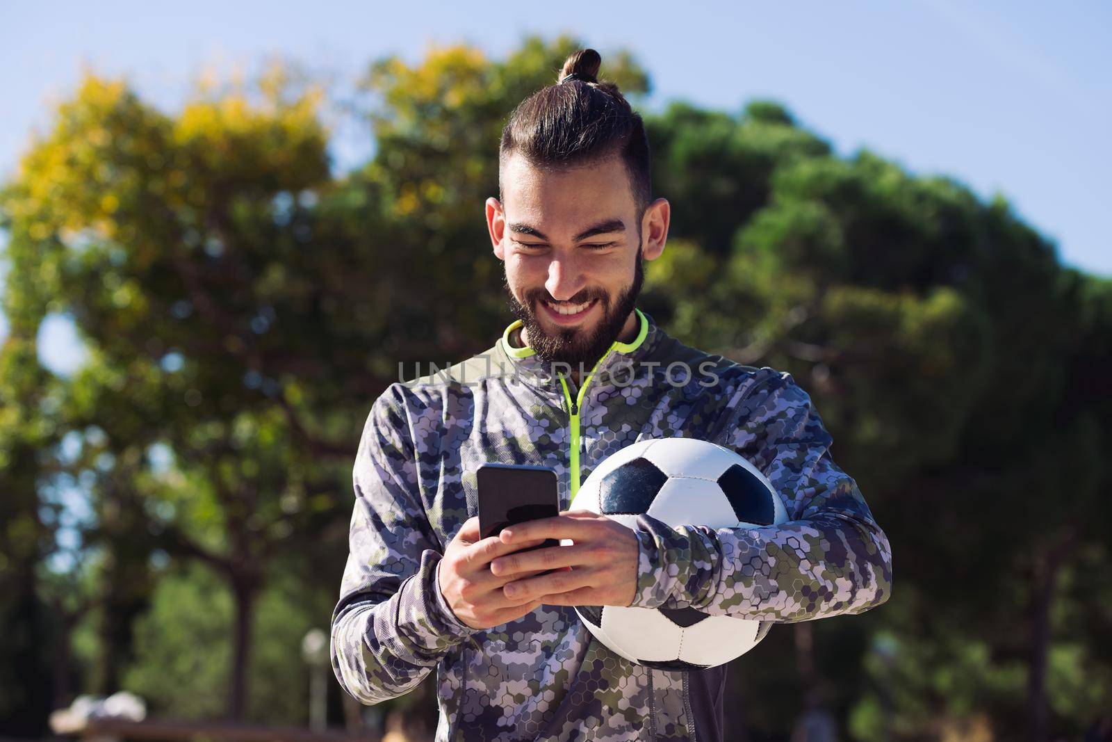 happy sportsman with a soccer ball smiles writing a message on his mobile phone, concept of technology and urban sport lifestyle in the city, copy space for text