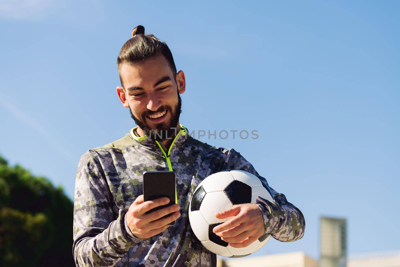 horizontal portrait of a happy sportsman with a soccer ball smiling looking his mobile phone, concept of technology and urban sport lifestyle in the city, copy space for text
