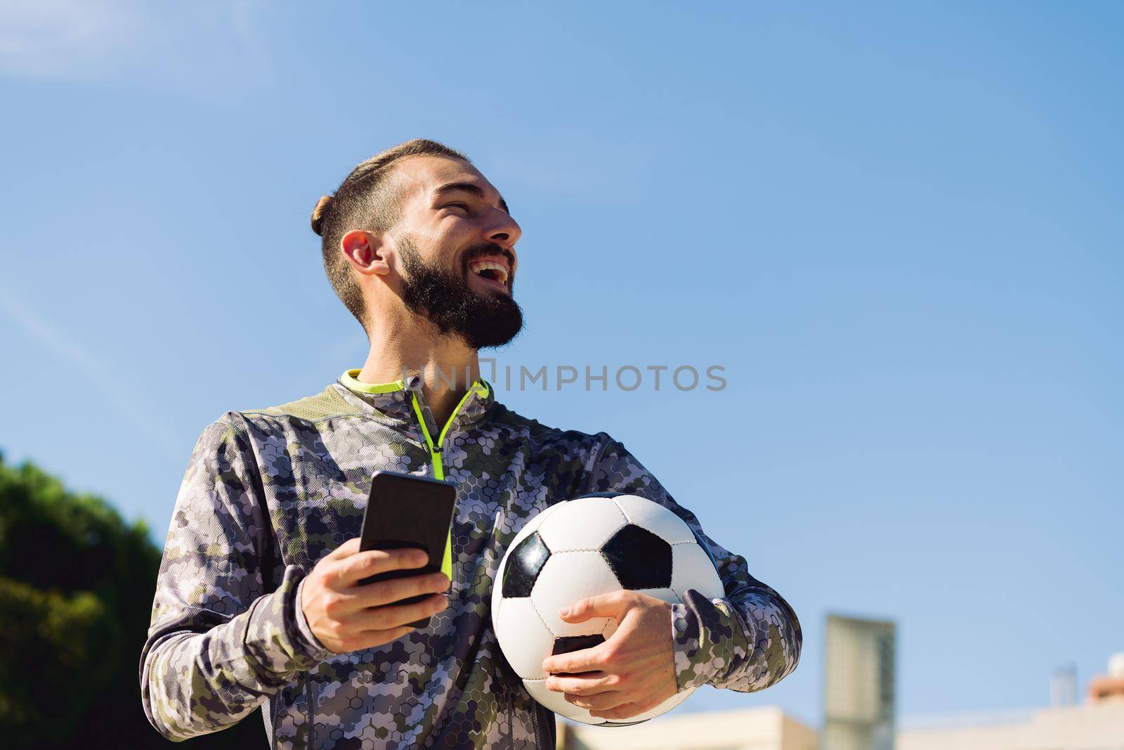 horizontal portrait of a happy sportsman with a soccer ball laughing with the phone in his hand, concept of technology and urban sport lifestyle in the city, copy space for text