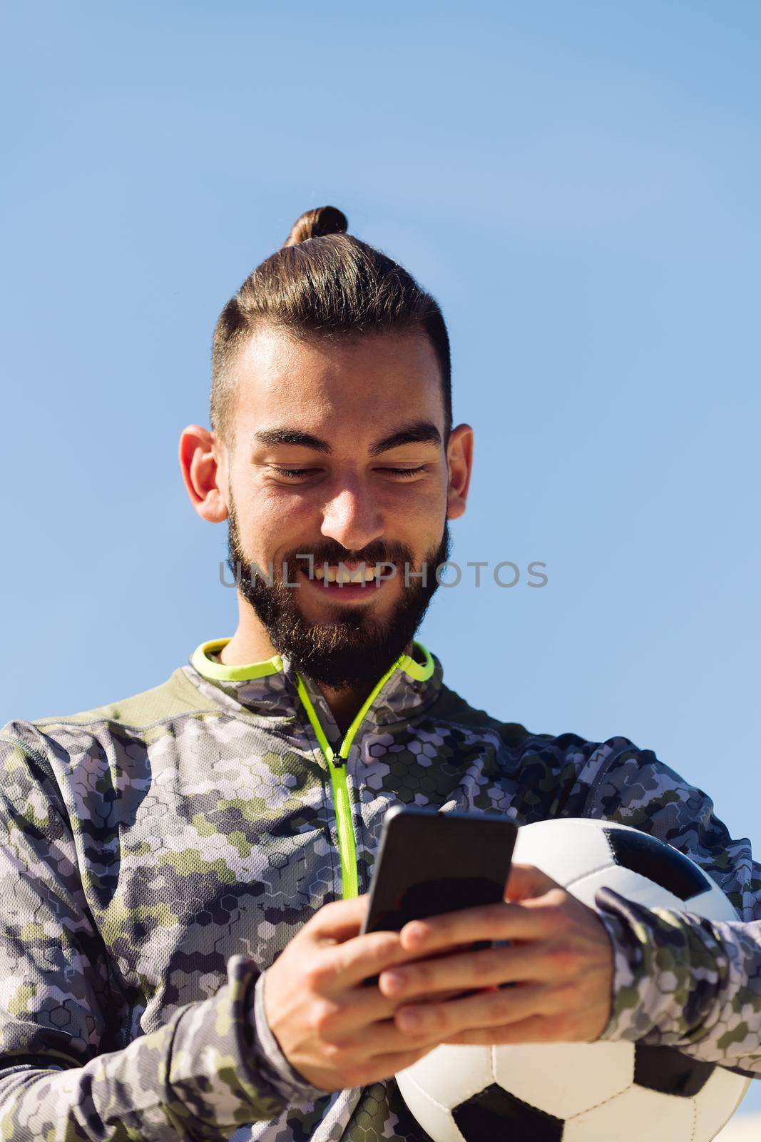 vertical portrait of a happy sportsman with a soccer ball laughing looking his cell phone, concept of technology and urban sport lifestyle in the city, copy space for text