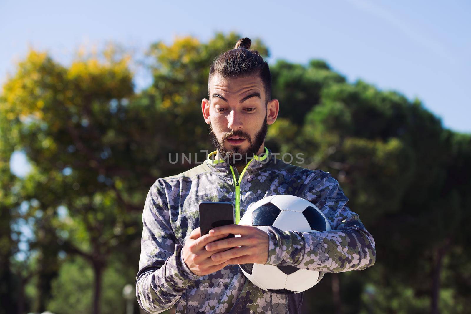 trendy sportsman with a football ball consulting his mobile phone with surprised expression, concept of technology and urban sport lifestyle in the city