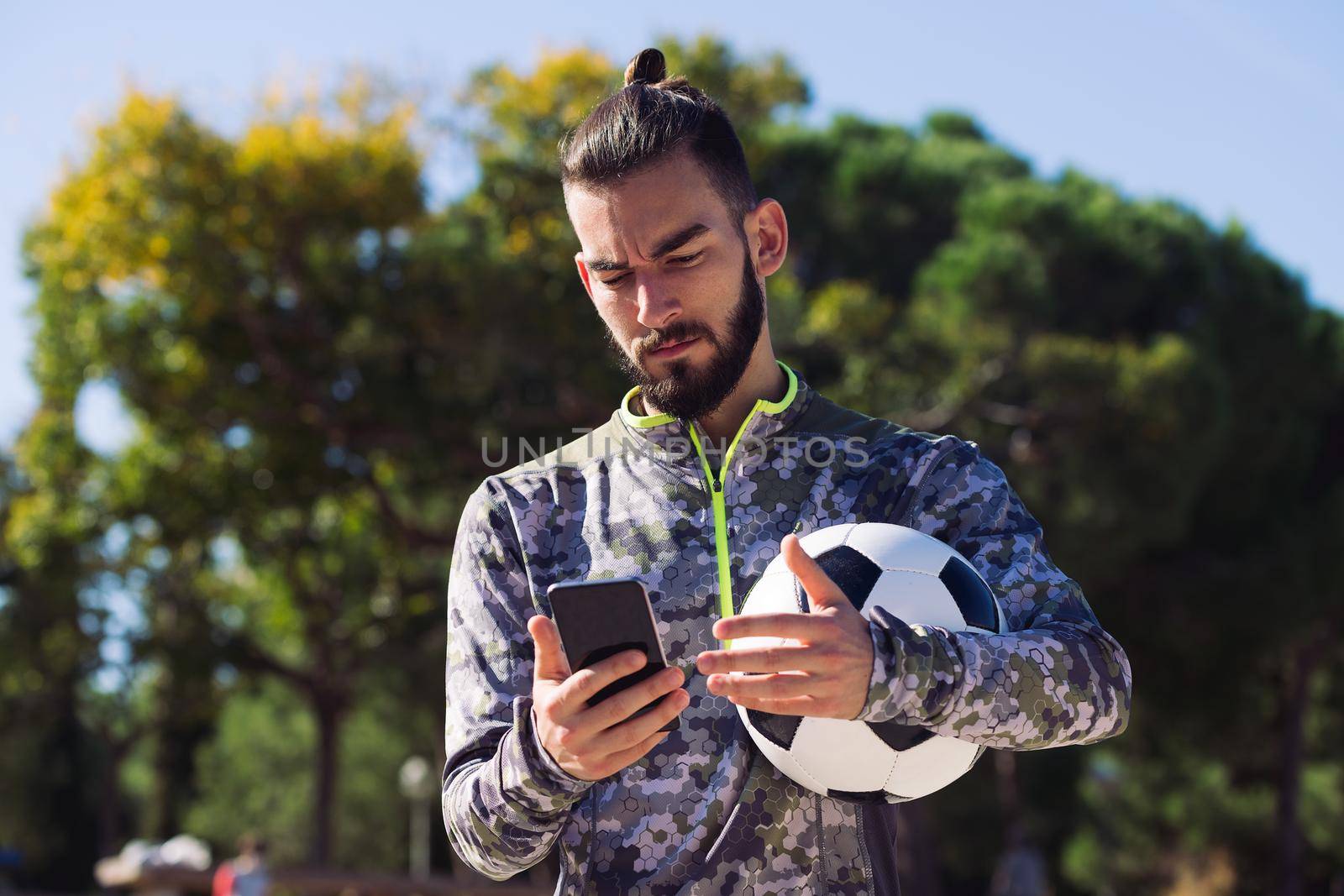 fashionable sportsman with a soccer ball consulting his mobile phone with confused expression due to an unexpected message, concept of technology and urban sport lifestyle in the city