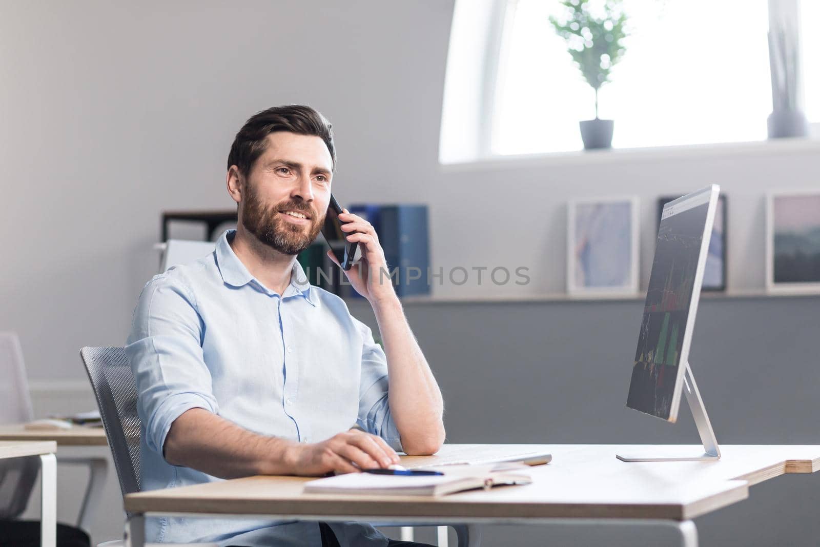 Young man with a beard, freelancer, manager, worker talking on a cell phone in the office, sitting at a computer desk, talking, waving