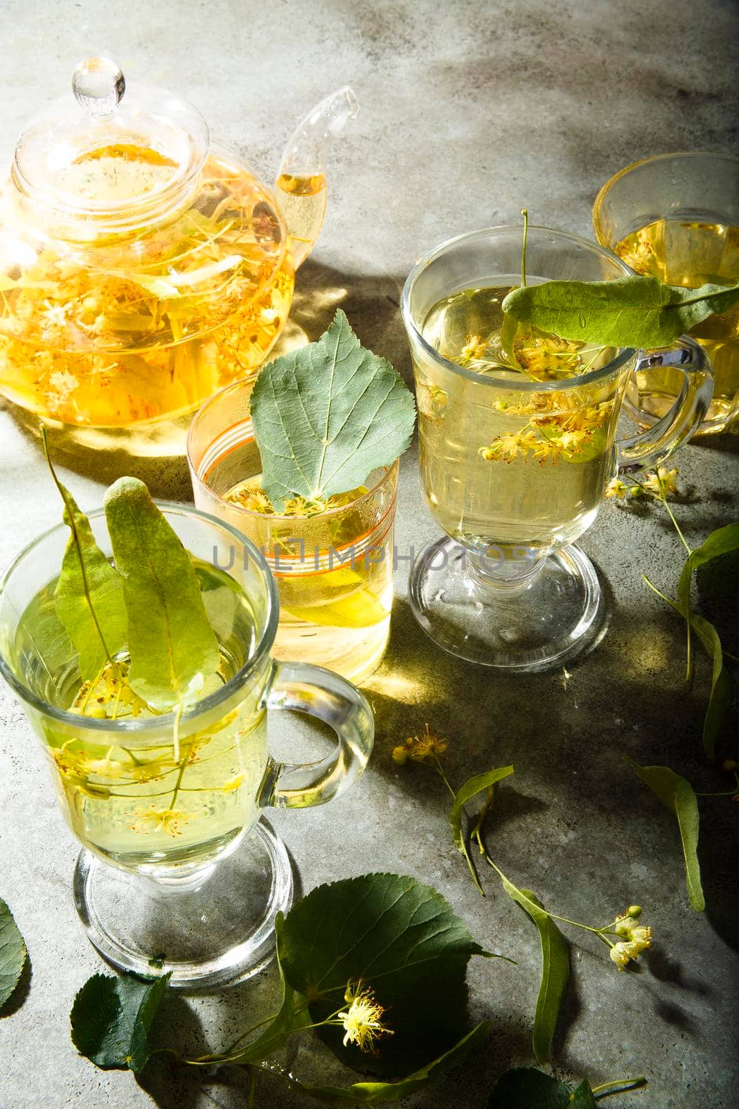 Glasses of freshly made linden tea and teapot on green table with leaves and flowers of linden tree, selective focus.