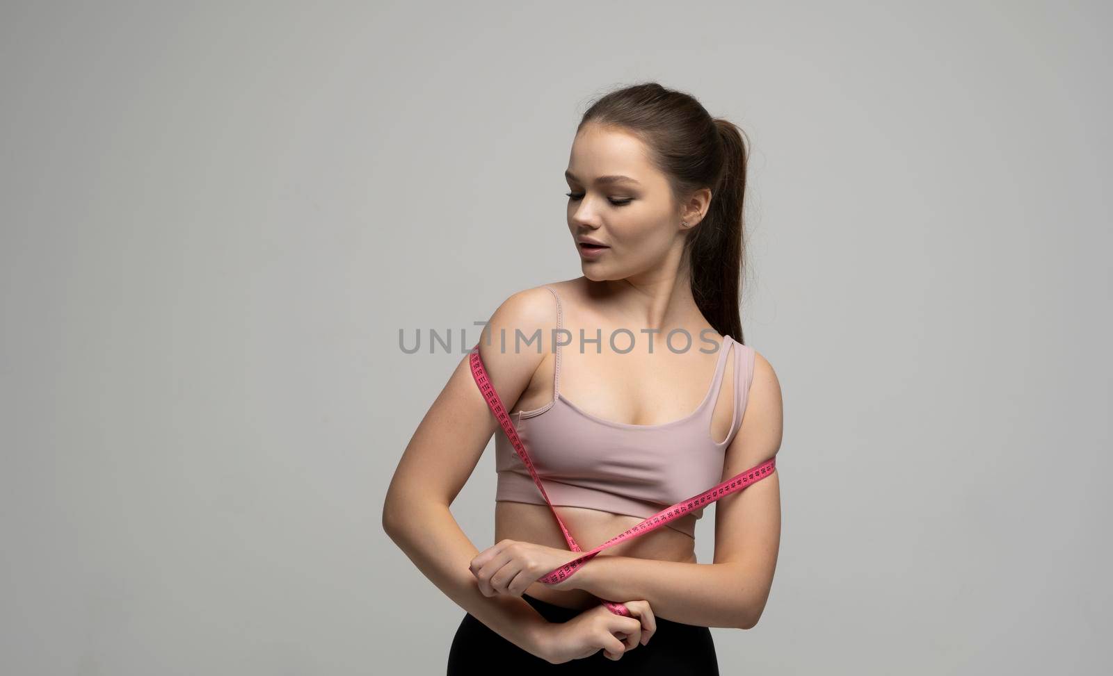 Young beautiful brunette woman in sport outfit sportswear holding a pink measuring tape. Weight loss and diet concepts. Health care and healthy nutrition. Perfect slim body. by vovsht