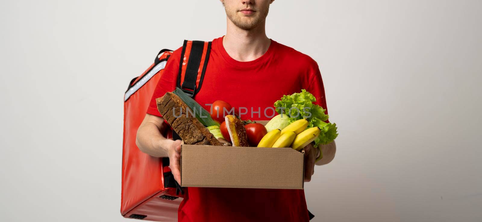 Crop picture of delivery man in red t-shirt carrying paper box with fruits and vegetables isolated over white background. Food delivery service. by vovsht