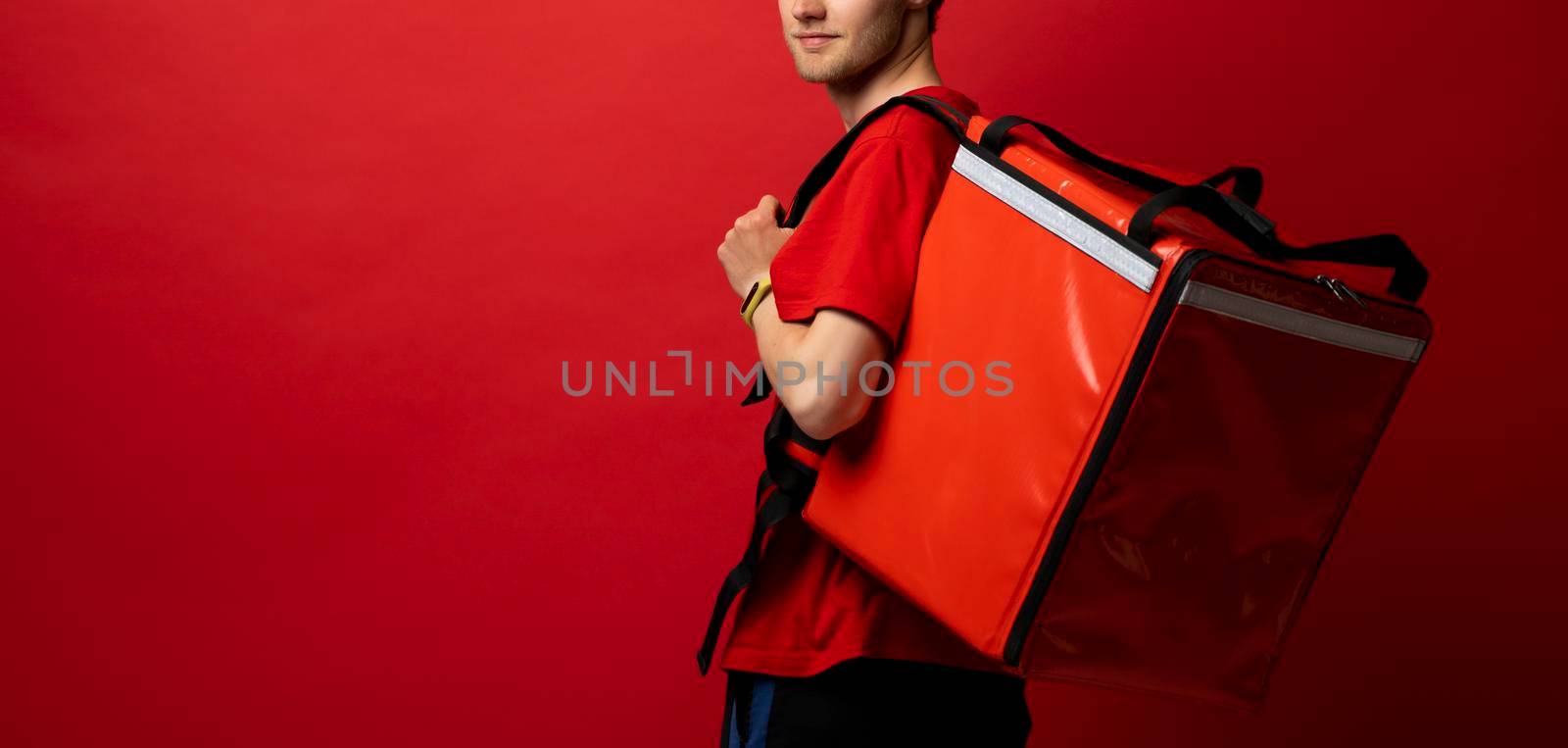 Portrait of young courier. Delivery man in uniform with thermo backpack isolated on red background. Fast home delivery. Online order