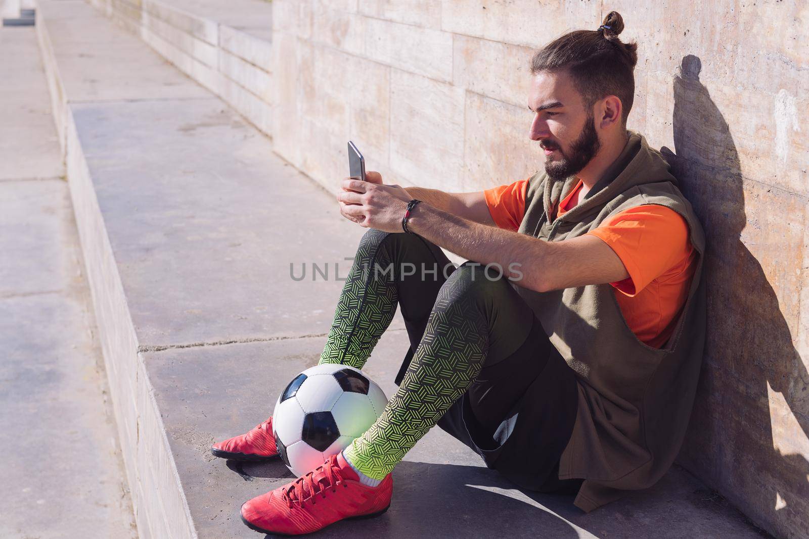fashionable football player rest sitting on the court consulting his mobile phone, concept of technology and urban sport lifestyle in the city, copy space for text