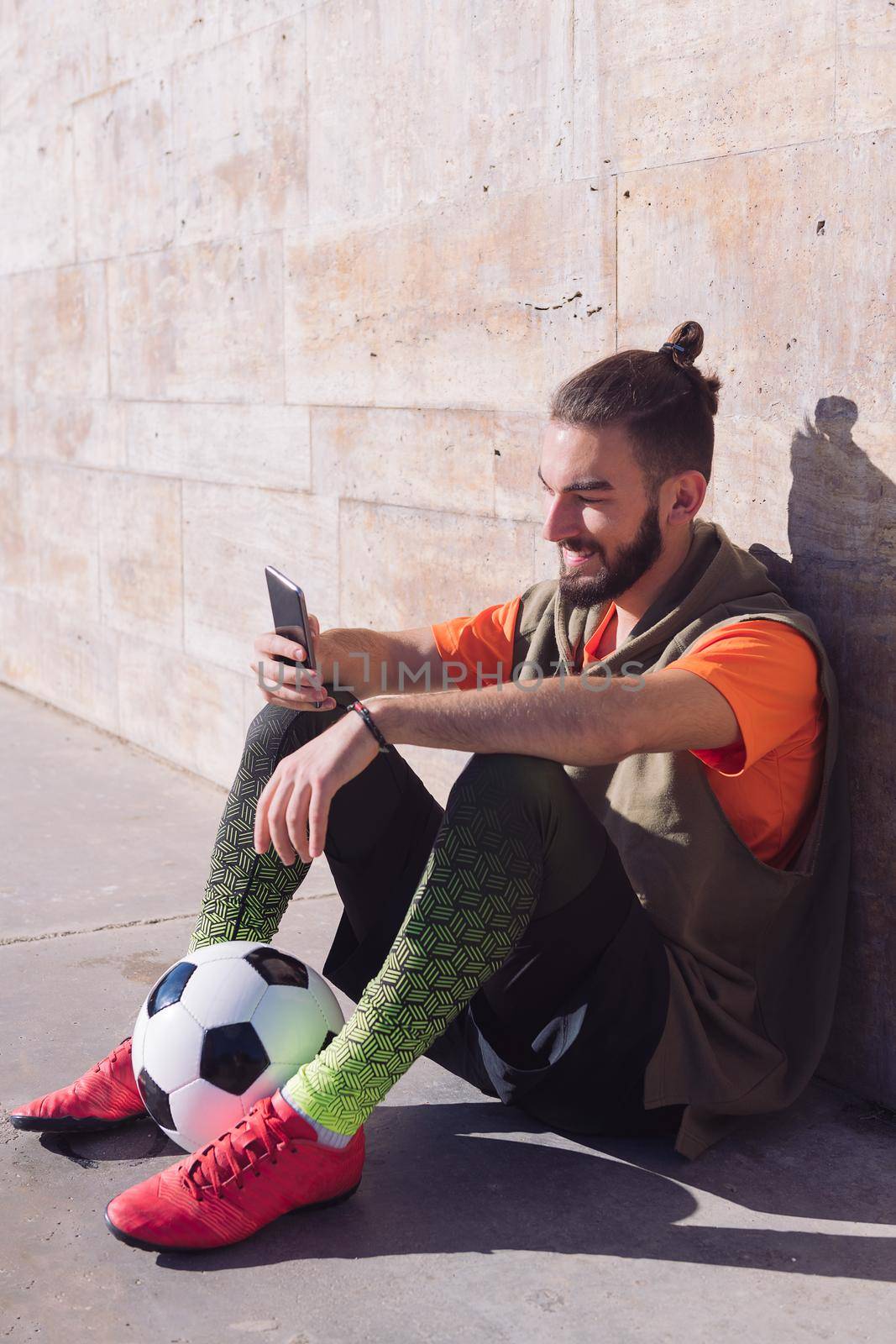 vertical photo of a fashionable soccer player rest sitting on the court consulting his mobile phone, concept of technology and urban sport lifestyle in the city, copy space for text