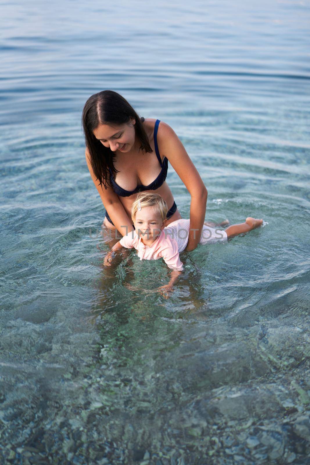 Mom holds a little girl by the stomach in the sea in shallows. High quality photo