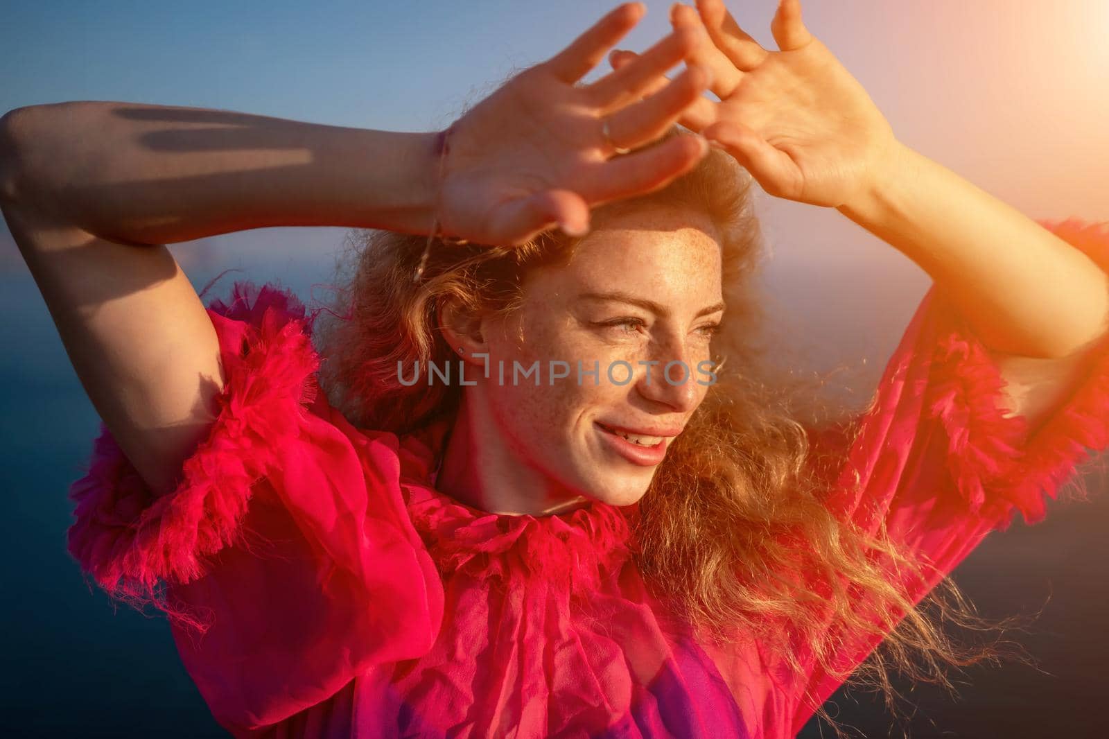 Close up shot of beautiful young caucasian woman with curly blond hair and freckles looking at camera and smiling. Cute woman portrait in a pink long dress posing on a volcanic rock high above the sea