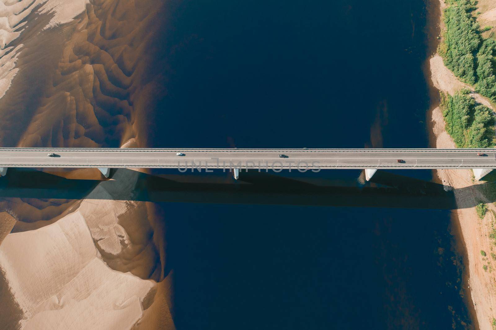 Aerial drone shot of cars driving on bridge over river and sandy riverside at sunset. Amazing aesthetic top view shot with beautiful coast with dune sands. Drone aerial bridge with car traffic