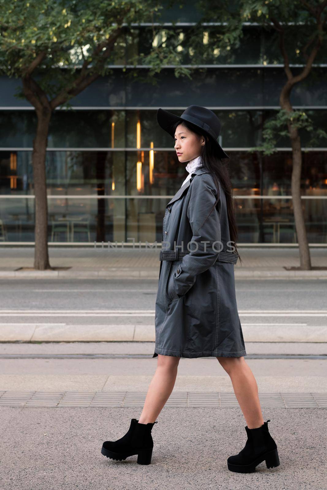 vertical photo of a classy asian woman walking in the city with hat and trench coat, concept of elegance and urban lifestyle, copy space for text