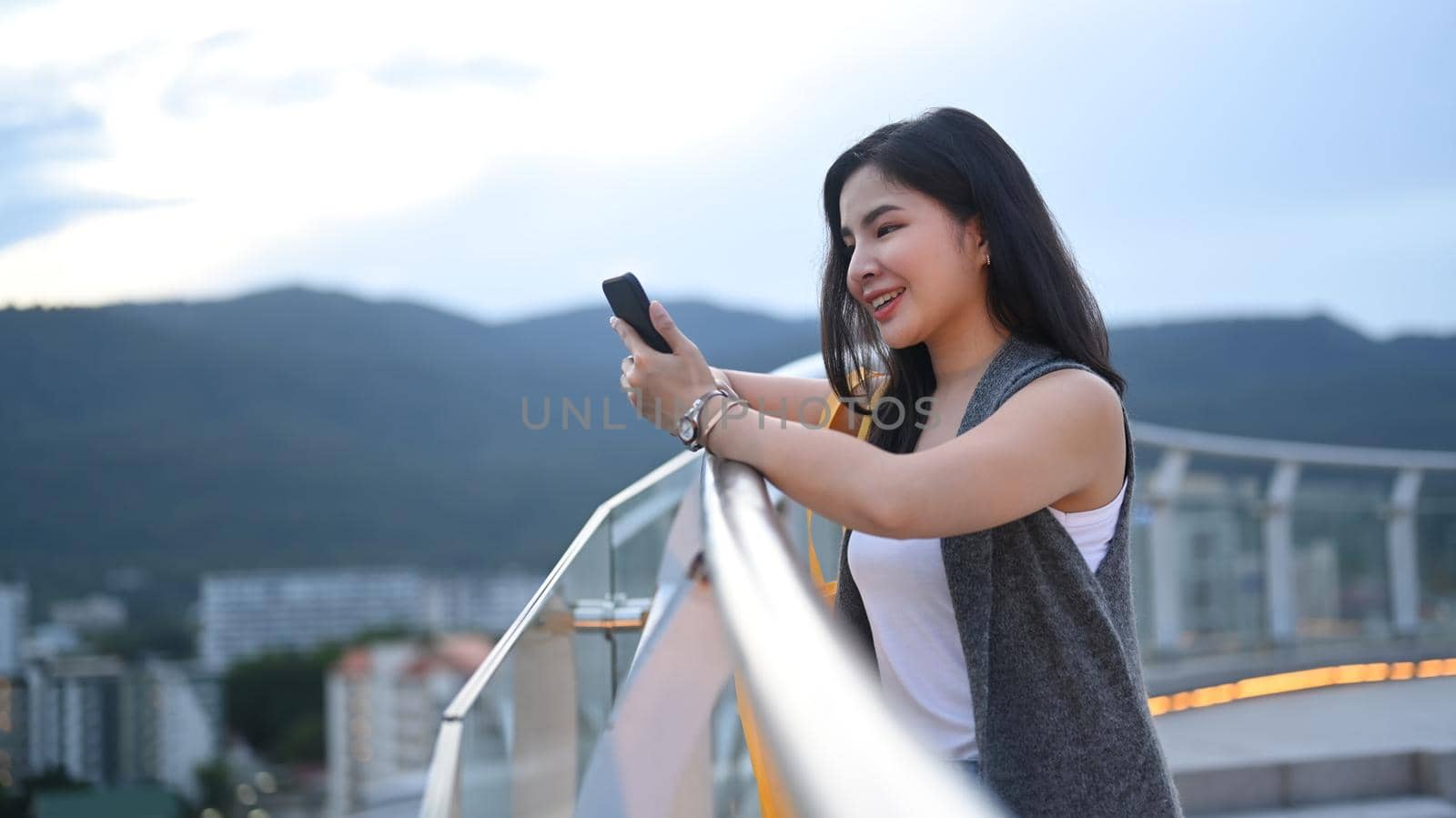 Charming young woman using mobile phone and standing on rooftop terrace with cityscape at sunset in background.