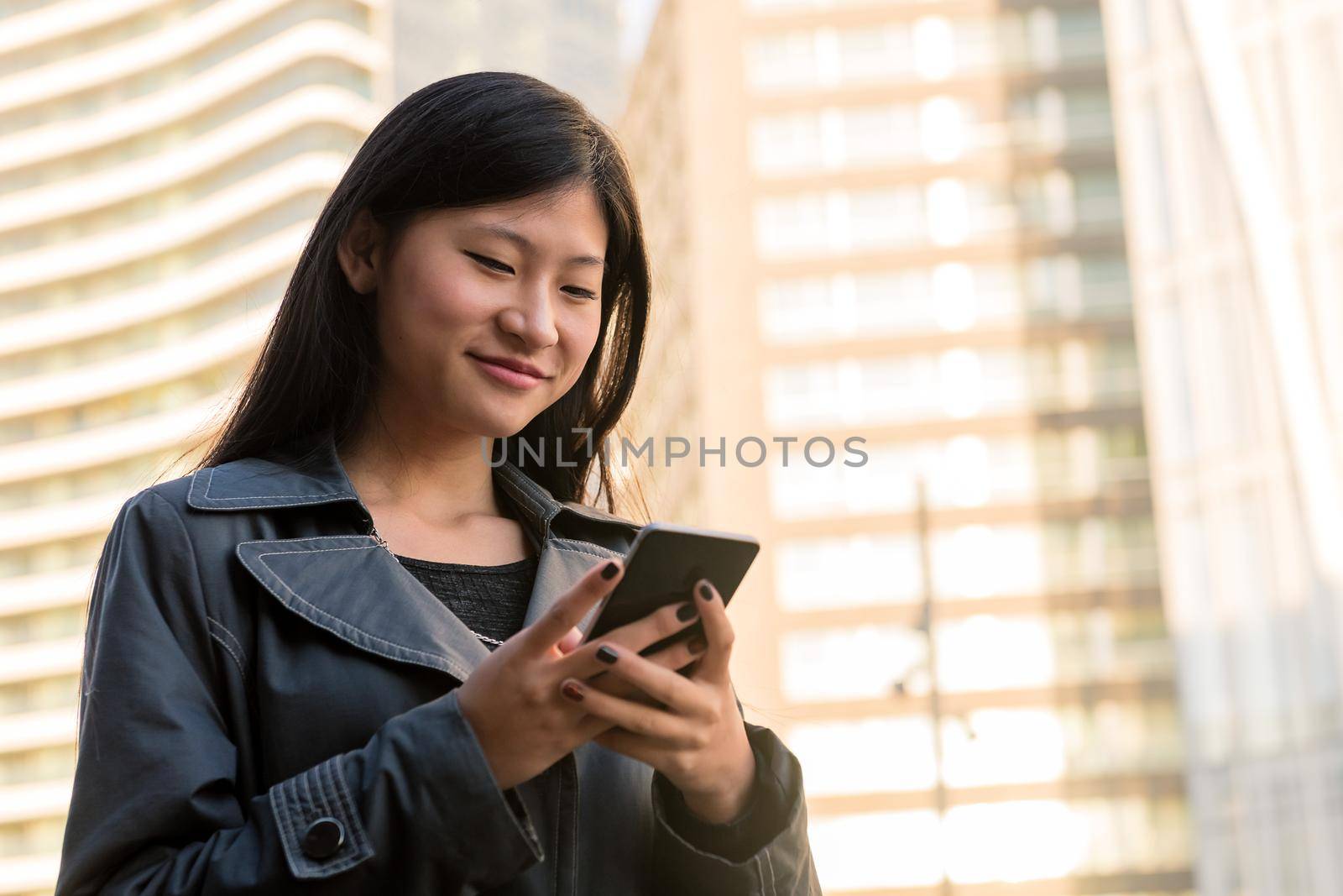 smiling young asian woman consulting her mobile phone in the city, technology and urban lifestyle concept, copy space for text