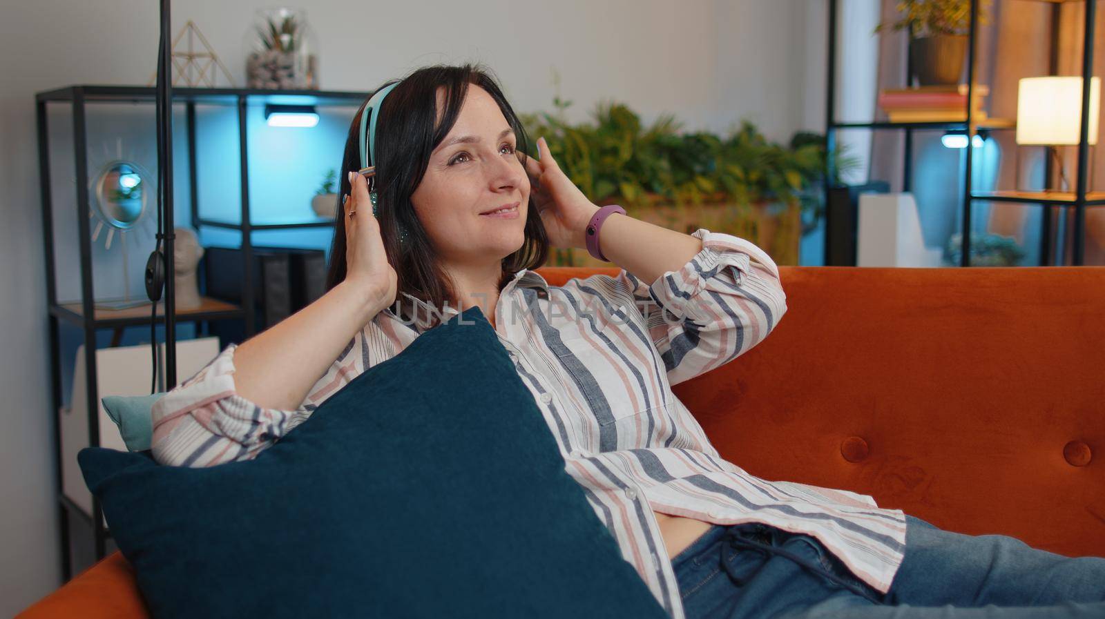 Happy relaxed overjoyed young woman in wireless headphones dancing on couch at home choosing listening favorite energetic disco rock n roll music in smartphone entertaining. People weekend activities