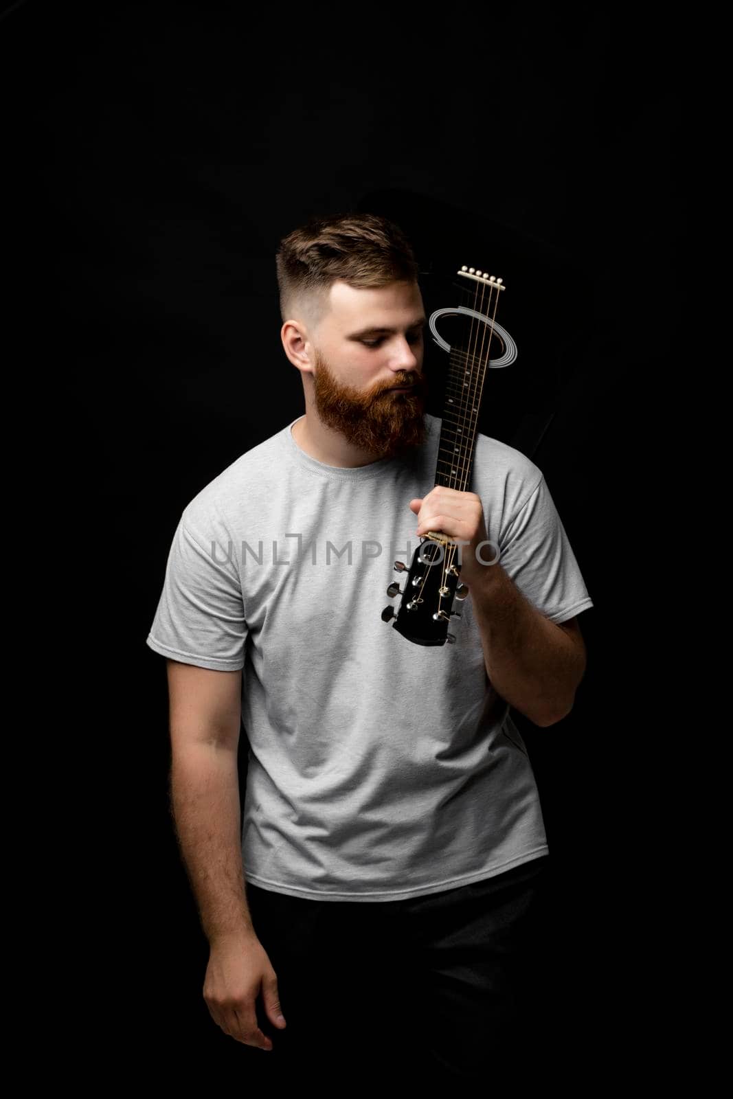 Brutal handsome bearded man in grey t-shirt with a guitar on a shoulder. Portrait of a male singer and guitarist