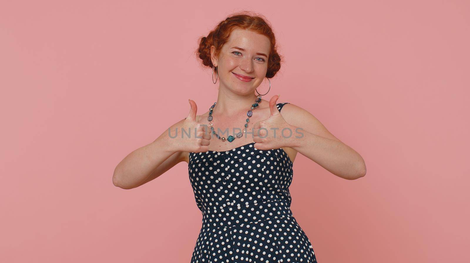 Redhead young woman in polkadot dress raises thumbs up agrees with something or gives positive reply recommends advertisement likes good. Lovely ginger girl isolated alone on pink studio background