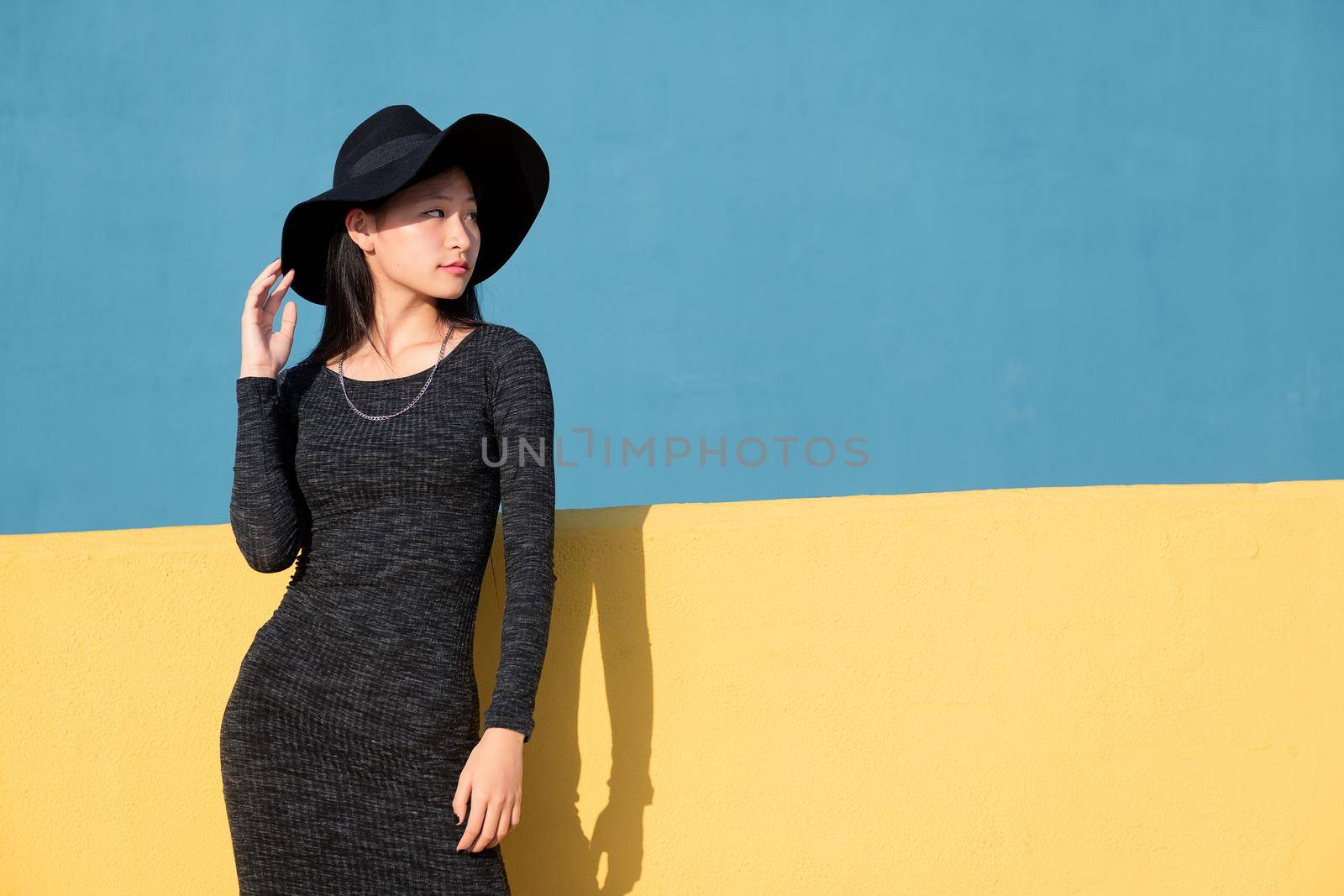 fashionable oriental young woman posing in a black tight dress and hat on a colorful yellow and blue background, concept of fashion and lifestyle, copyspace for text
