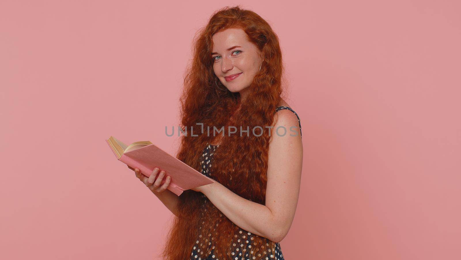 Redhead young woman reading funny interesting fairytale story book, leisure hobby, knowledge wisdom, education, learning, study, wow. Red hair ginger freckles girl isolated on pink studio background
