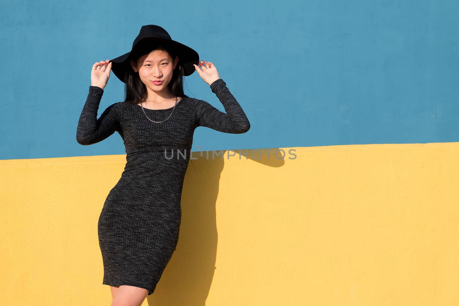 fashionable asian young woman posing in a black tight dress and hat on a colorful yellow and blue background, concept of fashion and lifestyle, copyspace for text