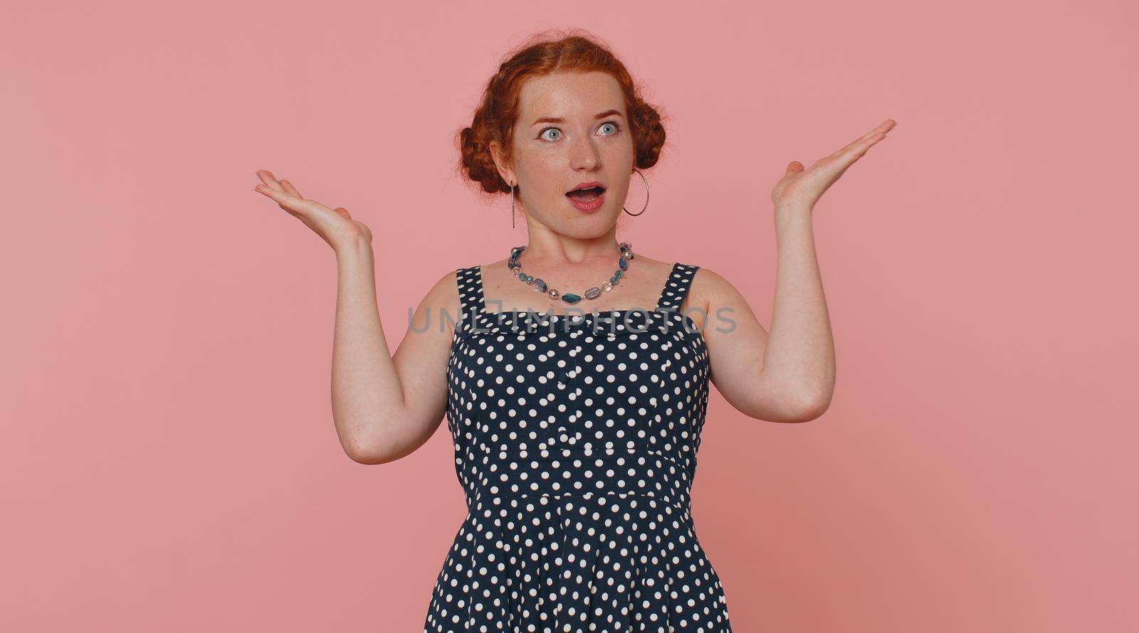 Excited amazed ginger woman in polkadot dress touching head, showing explosion, looking worried and shocked, professional burnout. Looking surprised wow girl isolated alone on pink studio background