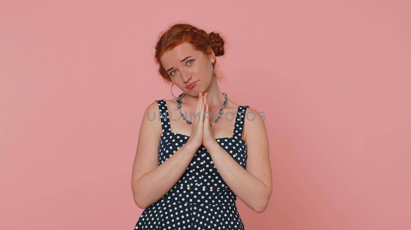 Please, God help. Young redhead woman praying, looking upward and making wish, asking with hopeful imploring expression, begging apology. Ginger girl with freckles isolated on pink studio background