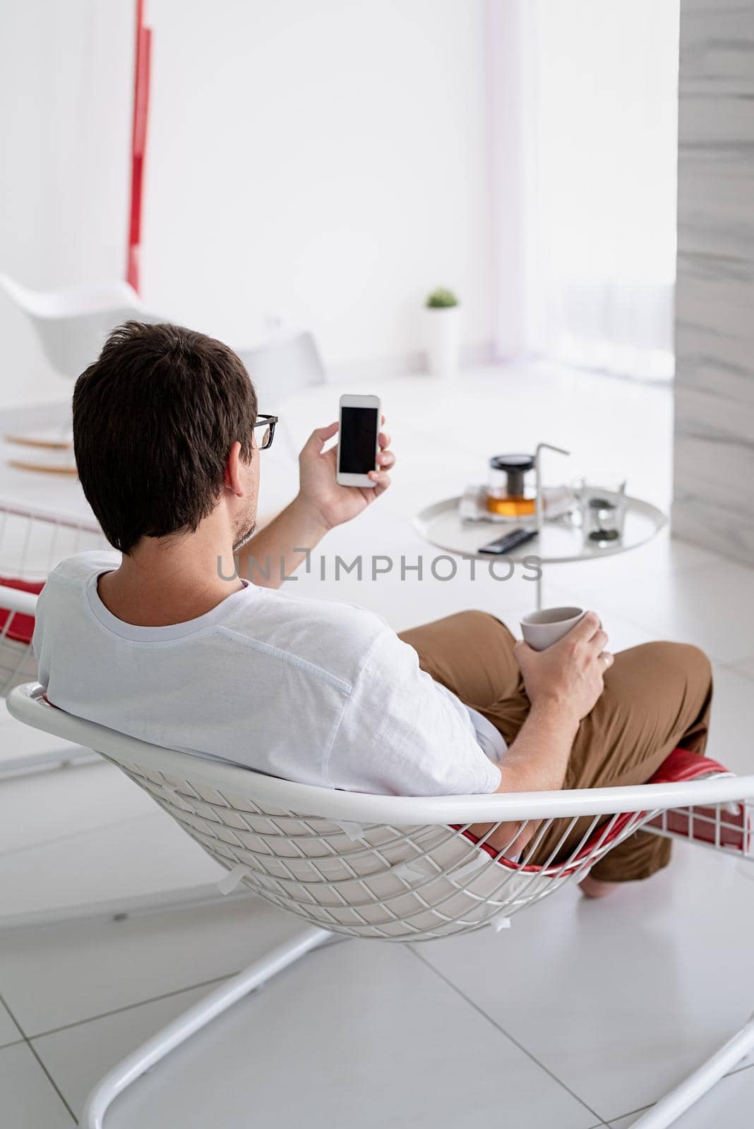 Leisure time. Man in white shirt sitting in a chair watching tv holding tea cup and phone