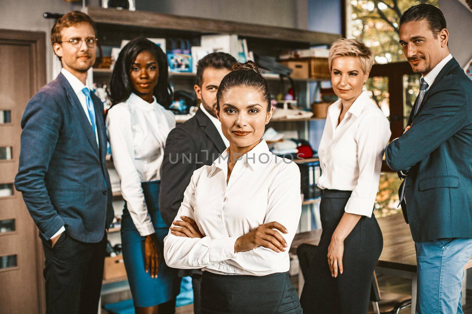 Multi-Ethnic Group Of Office Worker Posing Together, Business Colleagues In Formalwear Smiles Standing In Office, Selective Focus On Woman With Crossed Hands In Foreground, Toned Image