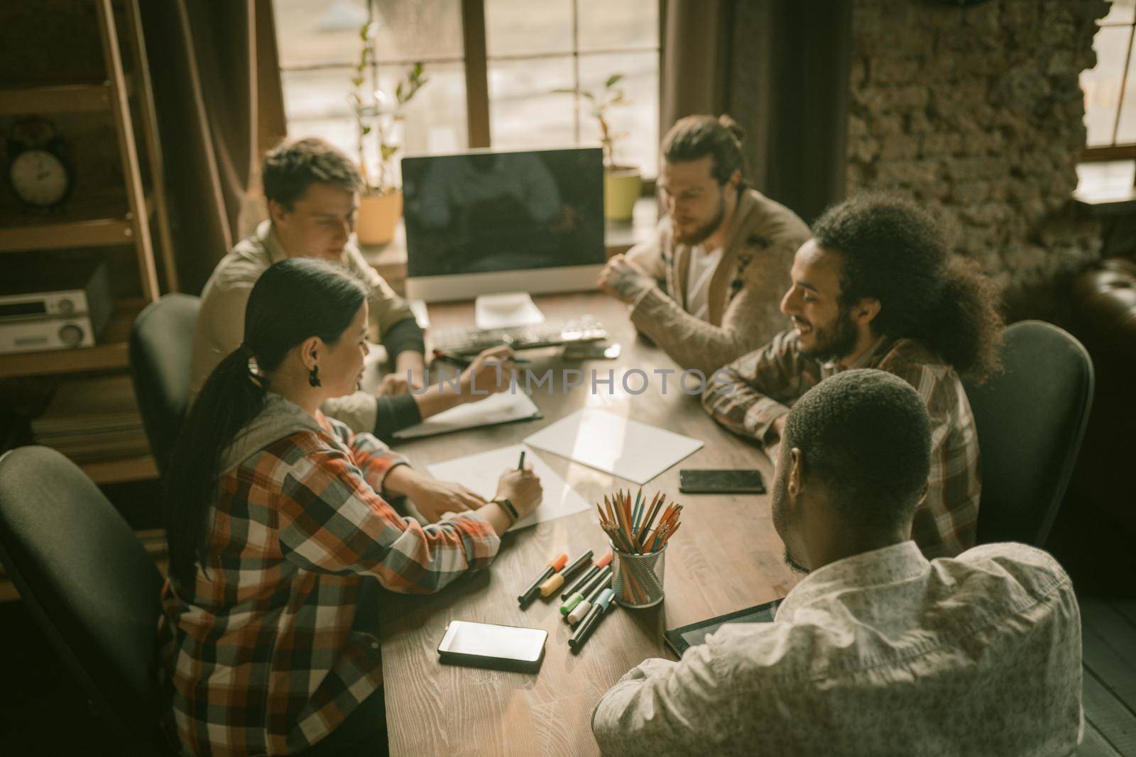 Multi-Ethnic Group Of Freelancers Or Students Doing Paper Work, Friends Are Planning Strategy Of New Startup Project Sitting At Large Wooden Table In Loft Interior Of Sunny Coworking Center