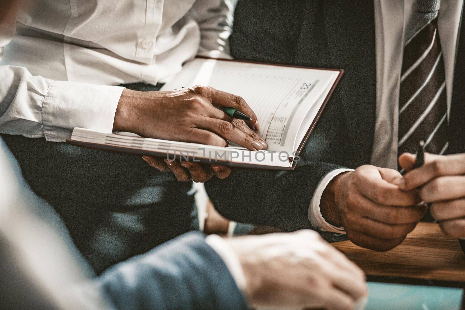 Female Office Manager Is Ready To Write In Notebook All Points Of Plan, While Business Partners Are Negotiating While Sitting At Office Table, Close Up Of Woman's Hand Holding Pen, Toned Image