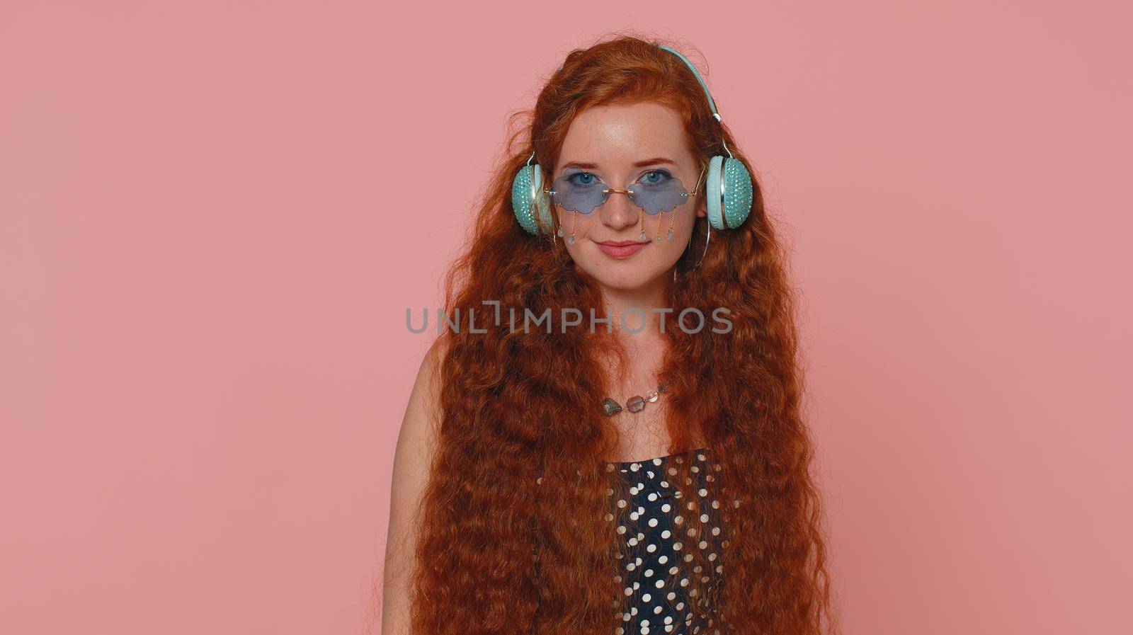 Young redhead woman in dress listening music on headphones, dancing disco fooling around having fun expressive gesticulating hands. Ginger girl with freckles isolated alone on pink studio background