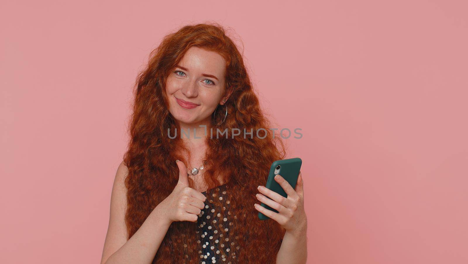 Beautiful red hair woman using mobile phone typing new post on web sms message, browsing, searching addiction of social networks. Young ginger girl studio shot isolated alone on pink wall background